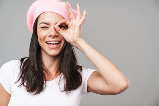 Portrait of a cute happy smiling young french woman in beret posing isolated over grey wall showing okay gesture.