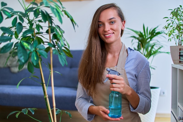 Portrait of cute happy young smiling attractive woman gardener in apron watering houseplants using spray bottle