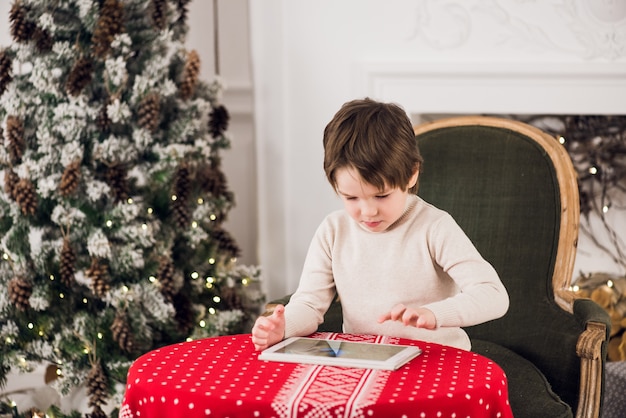 Portrait of cute kid boy sitting on green chair and playing with computer tablet