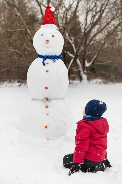 Foto ritratto di ragazzo carino seduto sulla neve e guardando il pupazzo di neve di bug nel parco invernale