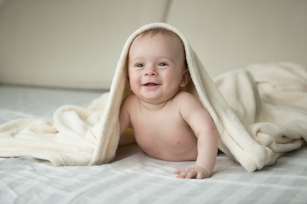 Portrait of cute laughing baby lying under blanket on bed