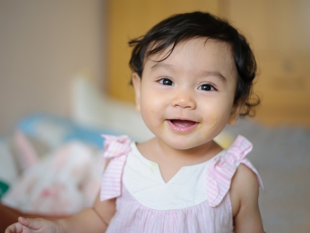 笑顔でベッドに座って、食べ物を口に塗ってカメラを見て幸せなかわいい小さなアジアのベイビーの肖像画。赤ちゃんの表現の概念
