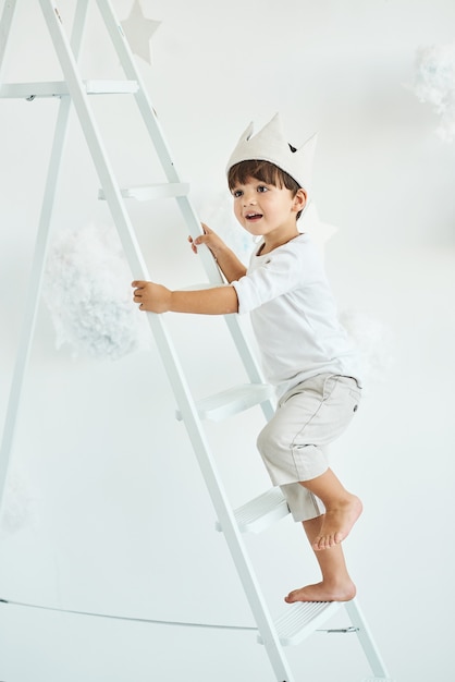 Portrait of a cute little boy in crown on white ladder among clouds on a white