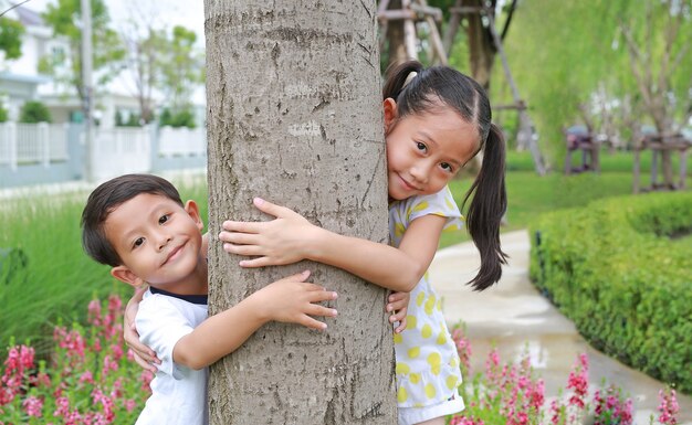 Portrait of cute little boy and girl child hugging with arms around tree trunk in the garden. Children enjoying nature life love, smiling in nature outdoors.