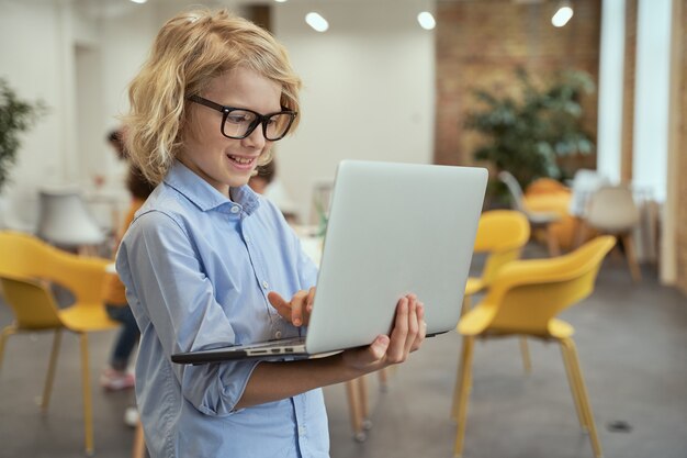 Ritratto di un ragazzino carino con gli occhiali che sorride mentre tiene e usa il laptop in piedi in a