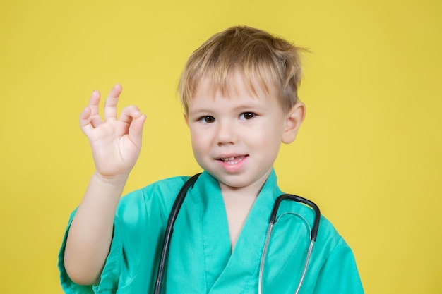 Portrait of cute little caucasian boy dressed in doctors green showing okay ok gesture on yellow background