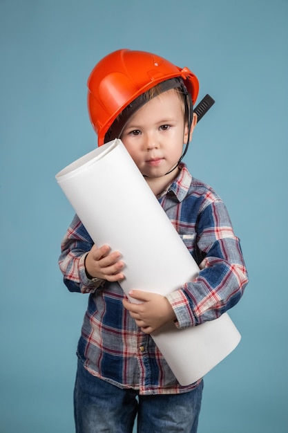 Portrait of cute little caucasian child builder in hardhats holding construction drawing against blue backfround. Concept of building a house for a happy family