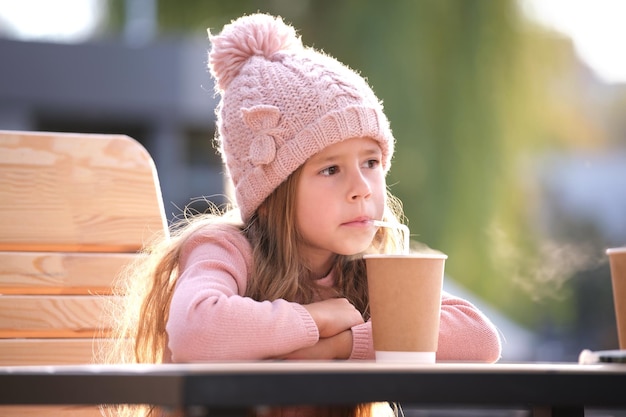 Portrait of cute little child girl in pink hat sitting alone at street cafe drinking tea from paper cup