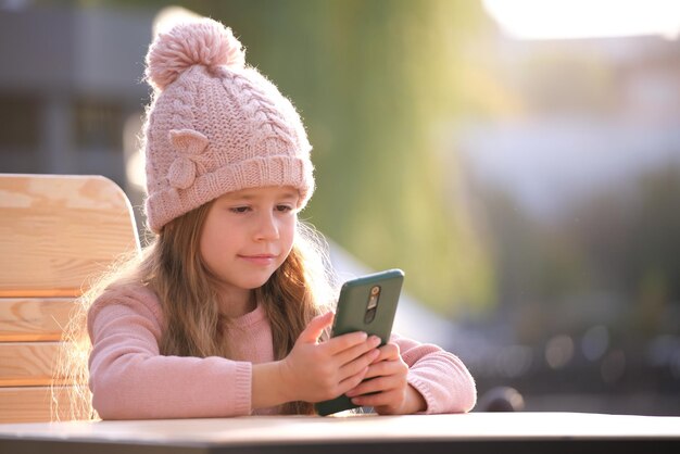 Portrait Of Cute Little Child Girl In Pink Hat Sitting Alone At Street Cafe Playing Game On Sellphone