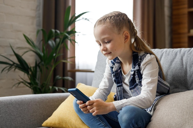 Portrait of cute little cute girl holding smartphone and\
resting at sofa at home choosing favorite music or cartoonstexting\
messagesbrowsing internetwatching video playing games on mobile\
phone