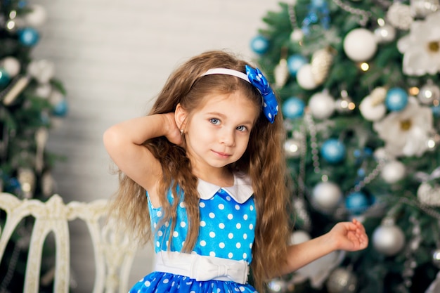 Portrait of a cute little girl in a blue dress posing, near the Christmas tree. Festive atmosphere.