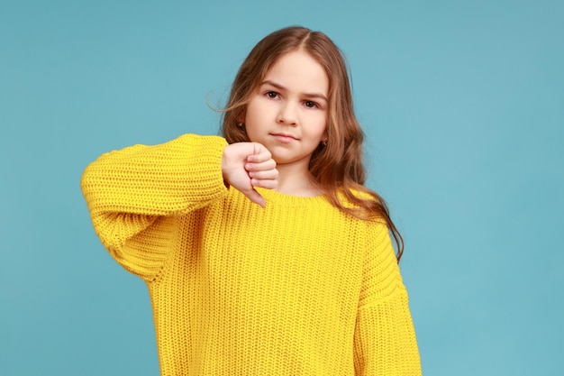 Portrait of cute little girl frowning and gesturing thumbs down, showing negative disapproval sign, wearing yellow casual style sweater. Indoor studio shot isolated on blue background.