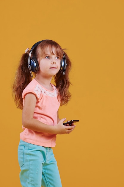 Portrait of cute little girl in headphones and with phone in the studio against yellow background