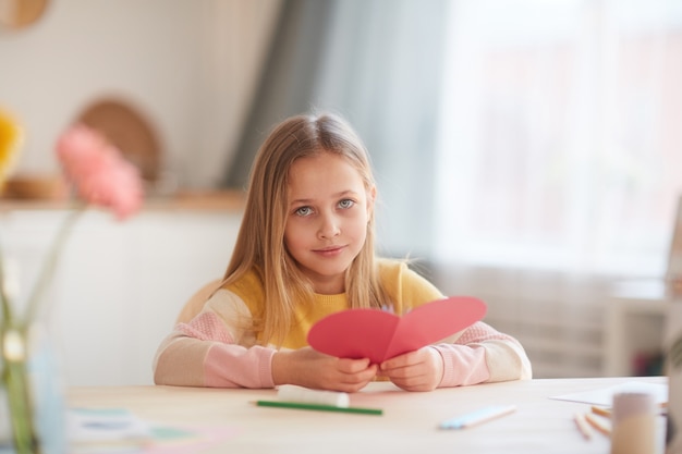 Portrait of cute little girl holding heart-shaped card while sitting at table in cozy home interior, copy space