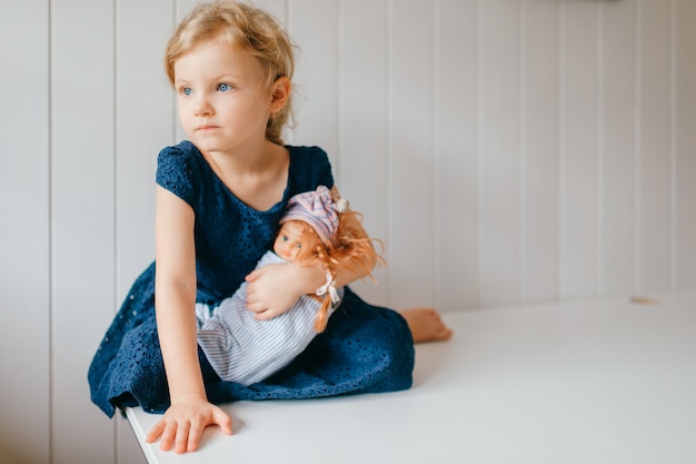 Portrait of cute little girl holds her lovely barbie, sits in bright baby room, looks aside