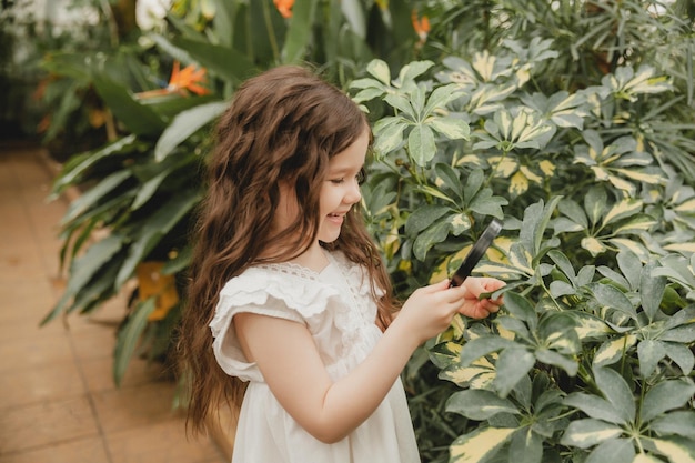 虫眼鏡を通して植物を見ているかわいい女の子の肖像画 虫眼鏡を持つ子供が庭で自然を研究します 初期開発の概念