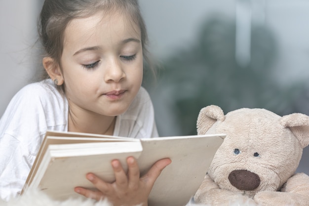 Portrait of a cute little girl reading a book at home, lying on the floor with her favorite toy.