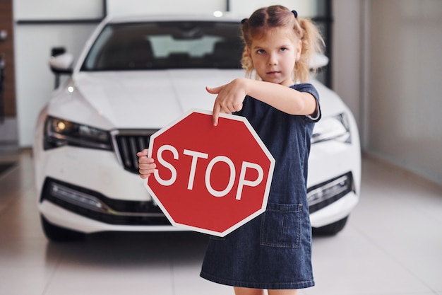 Portrait of cute little girl that holds road sign in hands in automobile salon.