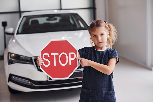 Portrait of cute little girl that holds road sign in hands in automobile salon.