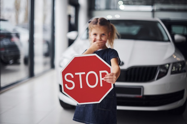 Portrait of cute little girl that holds road sign in hands in automobile salon.