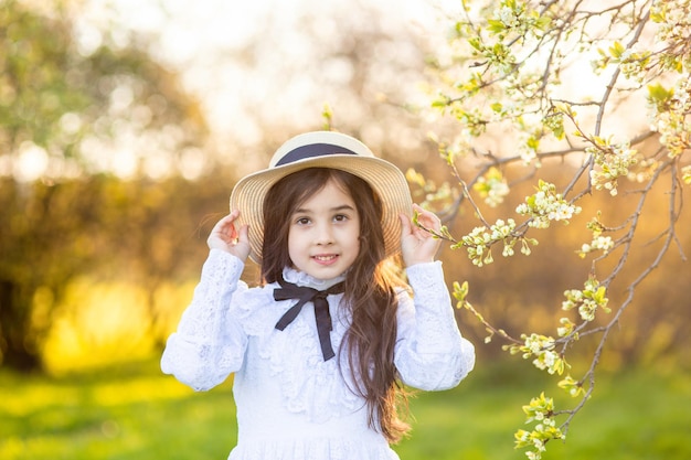 Portrait of cute little girl in a white dress and hat standing under flowering trees in spring