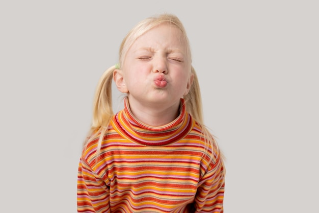 Portrait of a cute little girl with blond hair blowing a kiss to the camera with closed eyes isolated on gray