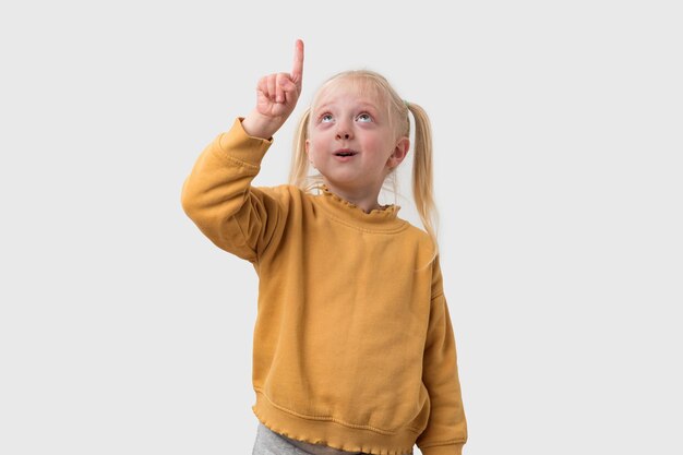 Portrait of a cute little girl with blond hair wearing a yellow sweater pointing up isolated on white background