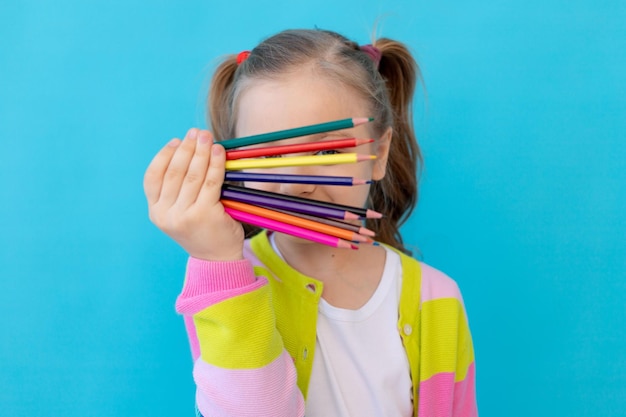 portrait of a cute little girl with colored pencils in a striped jacket. the concept of education and drawing. photo studio, blue background, place for text