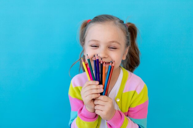 Портрет милой маленькой девочки с цветными карандашами в полосатой куртке Концепция образования и рисования Фотостудия синий фон место для текста