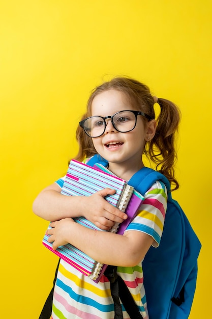 Portrait of a cute little girl with glasses in a striped T-shirt with notebooks and textbooks in her hands and a backpack. concept of education. photo studio, yellow background, space for text.