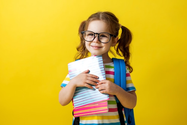 줄무늬 티셔츠에 안경을 쓴 귀여운 소녀의 초상화, 손에는 공책과 교과서, 배낭이 있습니다. 교육의 개념입니다. 사진 스튜디오, 노란색 배경, 텍스트를 위한 공간.
