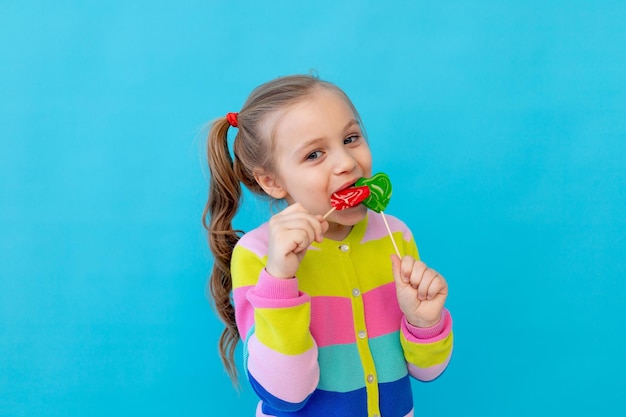 줄무늬 재킷에 막대 사탕을 입은 귀여운 소녀의 초상화 그 아이는 큰 막대 사탕을 먹고 핥습니다 과자와 사탕의 개념 텍스트를 위한 사진 스튜디오 파란색 배경 위치
