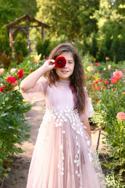 Foto un ritratto di una bambina carina con i capelli lunghi che indossa un abito lungo leggero in giardino. la ragazza si coprì gli occhi con un fiore