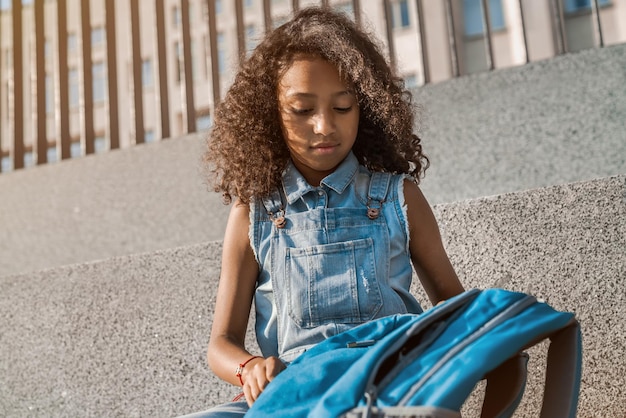Портрет милой маленькой девочки со школьным рюкзаком, сидящей на улице во дворе школы