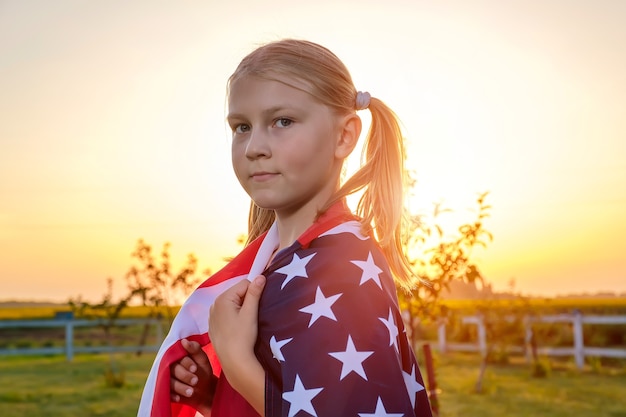 Ritratto di una bambina carina avvolta in una bandiera americana in piedi in un campo