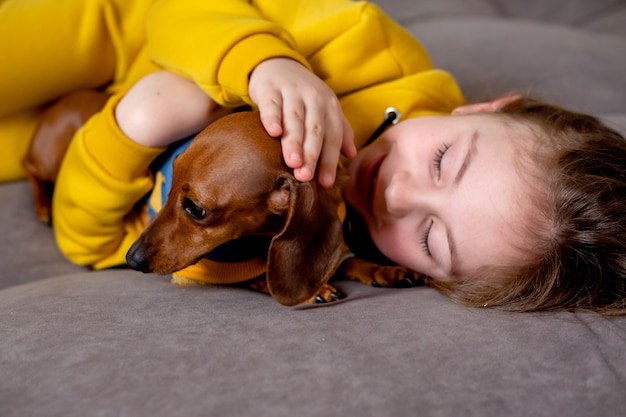 Portrait of cute little girl in yellow clothes lying in bed with a dwarf dachshund in blue jumpsuit