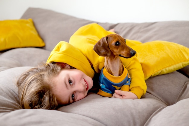 Portrait of cute little girl in yellow clothes lying in bed with a dwarf dachshund in blue jumpsuit