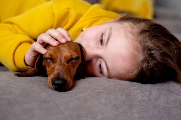 Portrait of cute little girl in yellow clothes lying in bed with a dwarf dachshund in blue jumpsuit