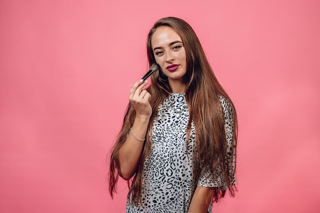 Portrait of cute lovely girl in casual outfit with the brush for make up holding in her hands, covering her face, looking at camera isolated on pink background. Skin care concept.