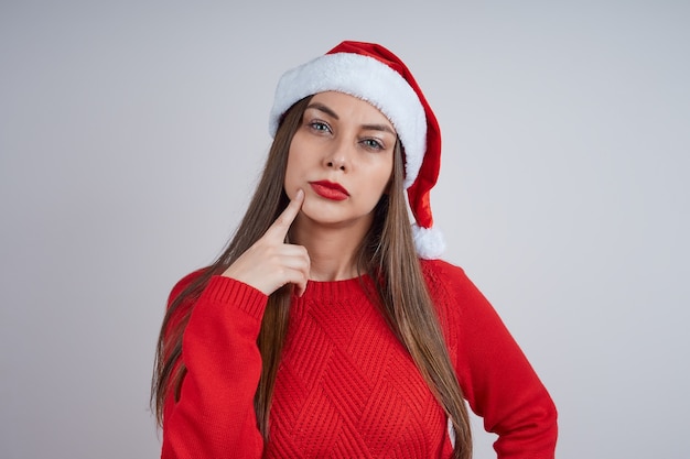Portrait of cute pensive woman in red sweater and santa hat on gray background.