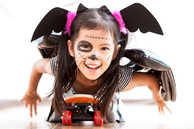 Portrait of cute playful girl with spooky make-up lying on skateboard at home during halloween