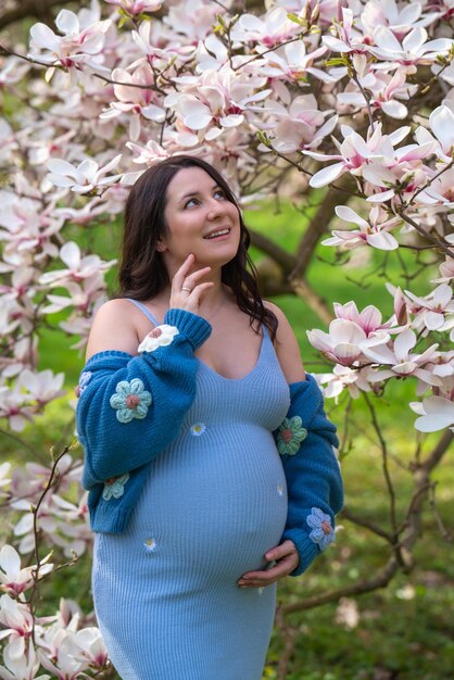 Photo portrait of a cute pregnant woman in the park dressed in a blue dress among magnoli flowers the expectant mother looks tenderly at a flowering tree pregnancy blooming magnolia