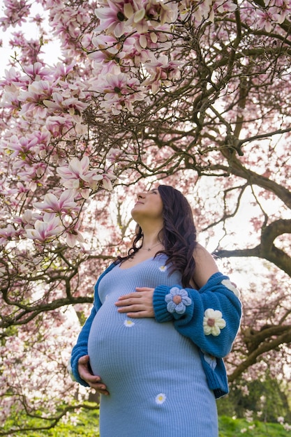 Photo portrait of a cute pregnant woman in the park dressed in a blue dress among magnoli flowers the expectant mother looks tenderly at a flowering tree view from below pregnancy