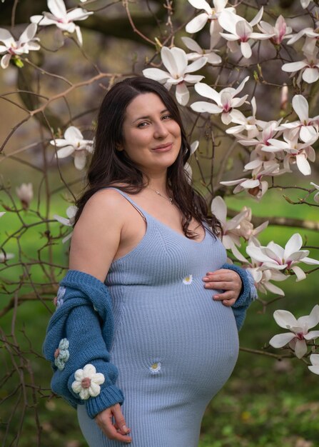 Photo portrait of a cute pregnant woman in the park dressed in a blue dress among magnoli flowers lady looking at camera closeup pregnancy waiting for the baby blooming magnolia