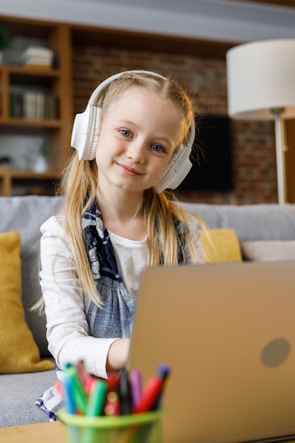 Portrait of cute primary school girl studying at home using laptop computer Smiling child wearing white earphones working on computer Distance learning Children home education