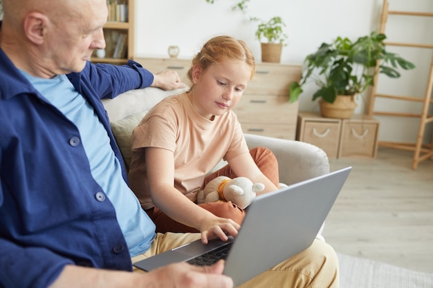 Portrait of cute red haired girl using laptop while enjoying time with grandfather in cozy home interior