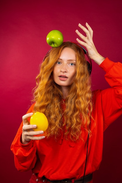 Portrait of a cute red-haired girl with long curly hair with freckles, with an orange, an apple and headphones. 