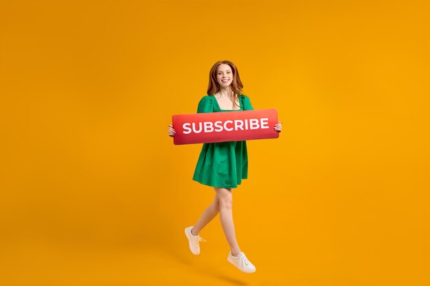 Portrait of cute redhead woman in green dress jumping, holding subscribe sign, ask appreciation by audience followers, popular forum. indoor studio shot isolated on yellow background