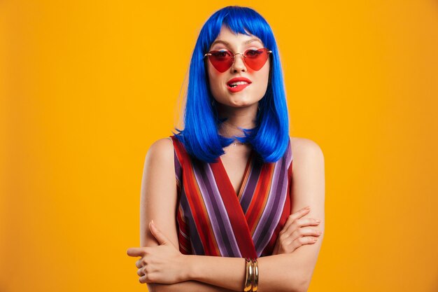 Portrait of cute seductive woman wearing blue wig and sunglasses looking and biting her lip isolated over yellow wall