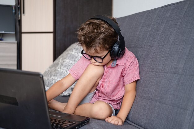 Portrait of cute serious caucasian boy using laptop and headphones while studying at home, remote education concept. Typing. Quarantine. Back to school concept.